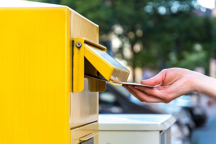 Gelber Postkasten, in den jemand einen Brief hineinwirft. 