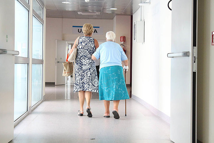 Alte Dame wird von jüngerer Dame einen Gang in einem Krankenhaus entlanggeführt.