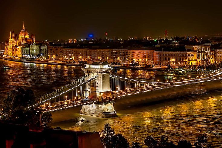 Budapest bei Nacht. Eine steinerne Brücke spannt sich über die Elbe. Im Hintergrund eine Kathedrale und repräsentative Bauten.