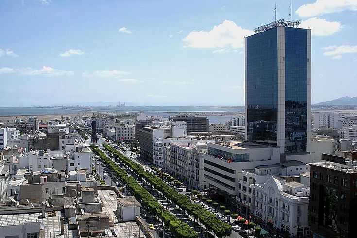 Bild der tunesischen Hauptstadt Tunis. Das Meer ist im Hintergrund erkennbar. Eine mit Bäumen gesäumte Hauptstraße, modern anmutender Büroturm, Hafenanlagen weiter hinten.