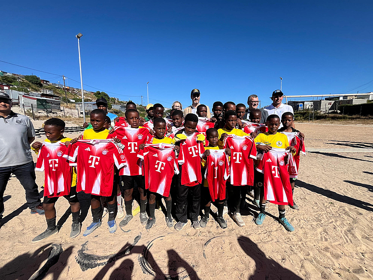 Ein große Gruppe von kleinen jungen in Fusßballtrickots stehtl am Strand. Im Hintergrund steht Dr. Markus Söder und Florian Herrmann.
