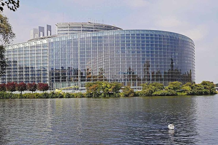 Das runde, gläserne Gebäude des EU-Parlaments an einem Wasser.