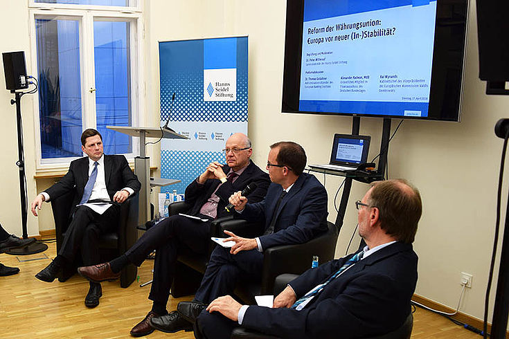 Vier Herren in Ledersesseln die gedankenvoll zuhören. Grave Stimmung. Im Hintergrund HSS-Stellwand und Bildschirm mit Präsentation.