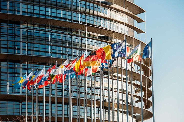 Ein großes, rundes Gebäude aus Glas und Stahl mit den Flaggen der EU-Mitgliedsstaaten davor, die im Winde wehen.