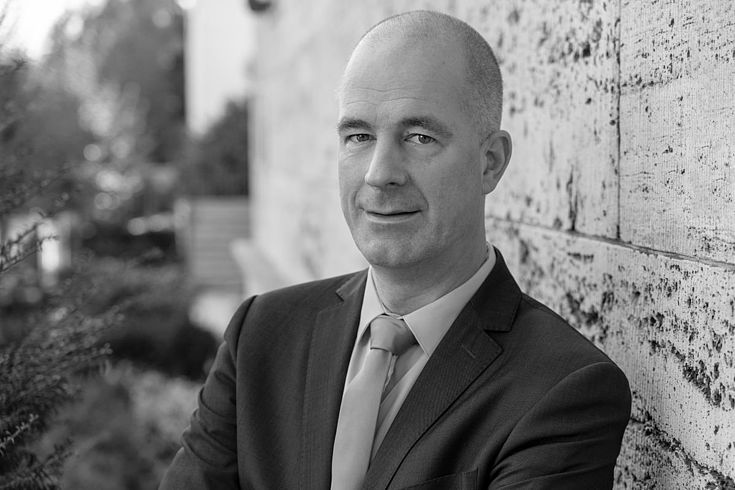 Ein Mann im Anzug lehnt an einer Mauer und blickt ernst aber freundlich in die Kamera.