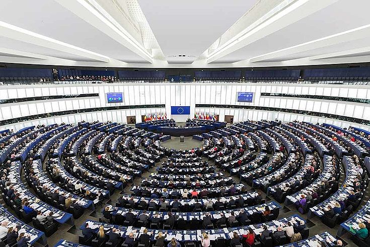 Der Plenarsaal des EU-Parlaments in Straßburg. Ein riesiger Raum, kreisrund mit hoher Decke und sternförmig auf einen nach hinten versetzten Mittelpunkt zulaufende Gänge zwischen dichten Tischreihen, die alle kreisförmig angeordnet sind.