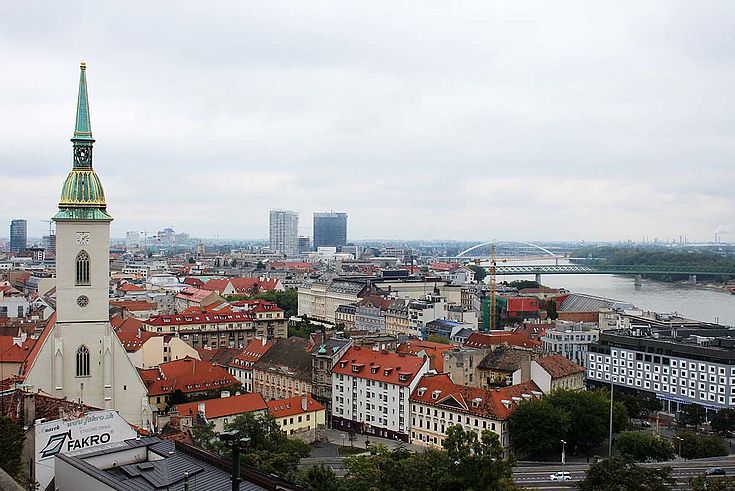 Blick auf Bratislava wohl von einem hohen Turm aus. Im Vordergrund ein spitz zulaufender Kirchturm. Rechts im Bild ein breiter Fluss, der von Brücken überspannt wird. 