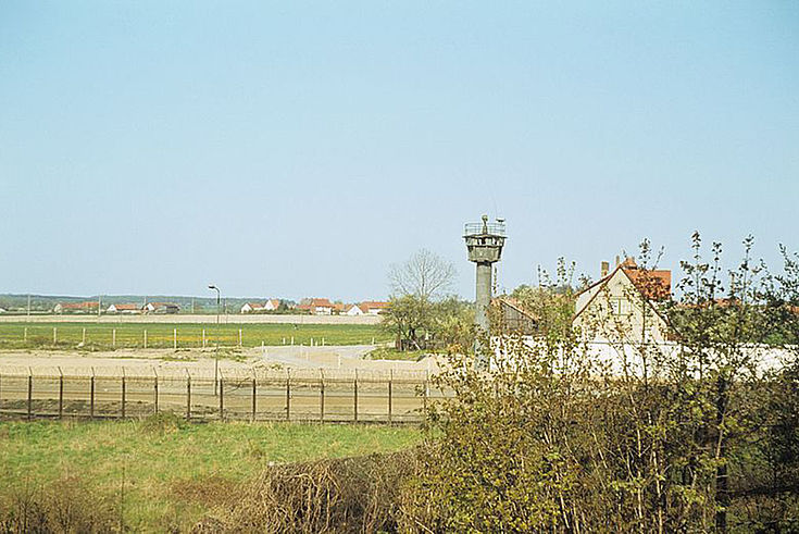 Blick auf die Grenze zwischen der BRD und der DDR. Hinter dem Grenzzaun erhebt sich ein Wachturm. Im Hintergrund ist ein Dorf zu sehen.