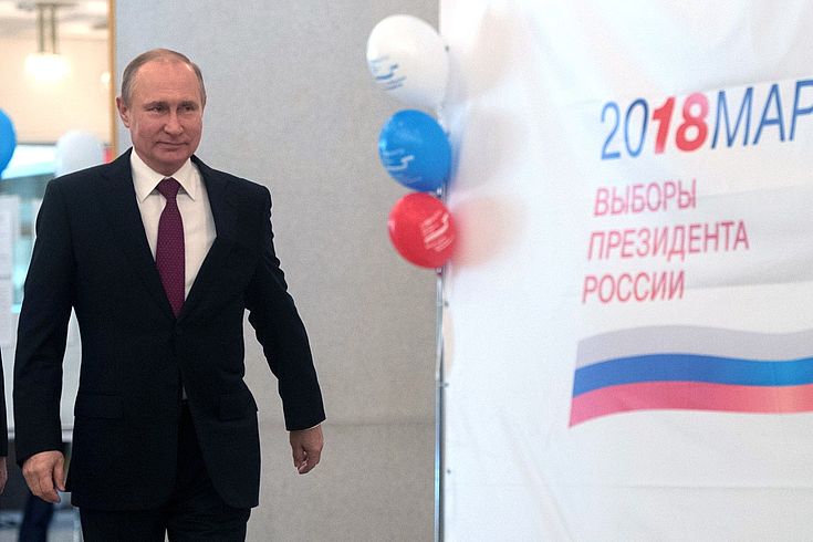 Putin schreitet forsch nach vorn. Daneben ein Wahlplakat.