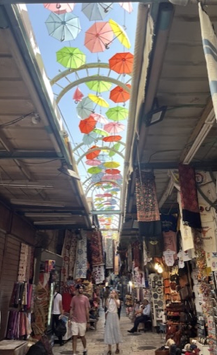 Eine Gasse in der Altstadt Jerusalems mit Sonnenschirmen. Links und rechts haben Händler ihre Shops, in denen sie Gewürze, Kleidung und Souvenirs verkaufen.