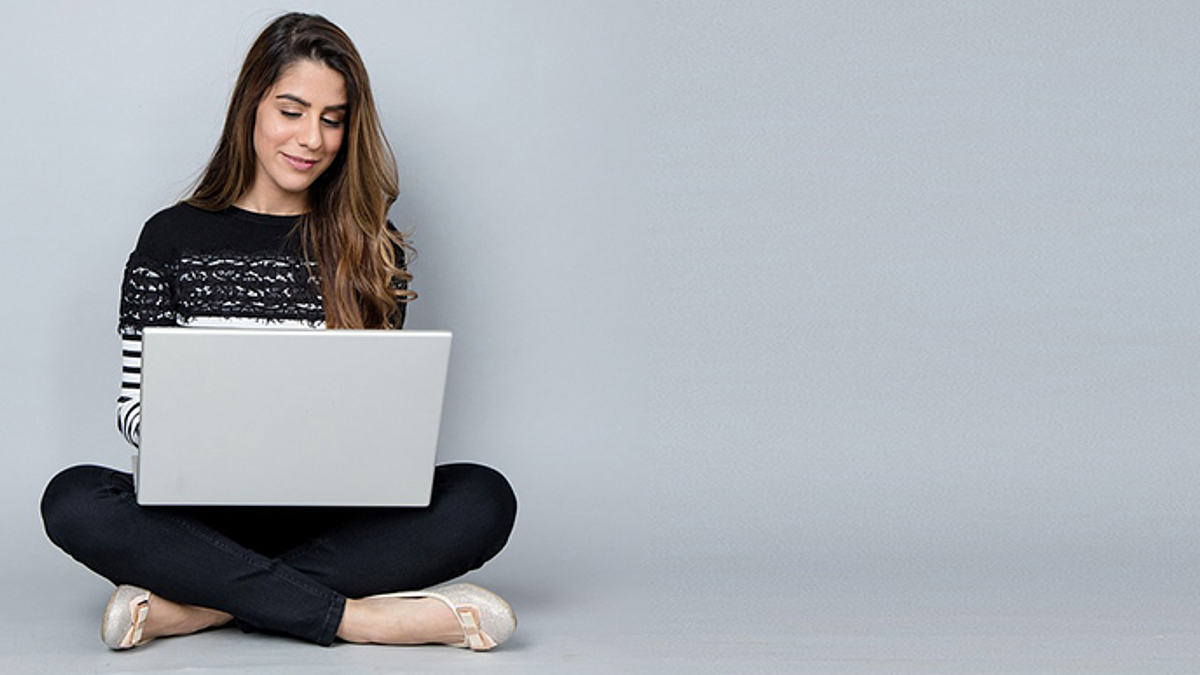 Frau sitzt in bequemen Klamotten auf dem Zimmerboden an der Wand und surft entspannt lächelnd auf ihrem Laptop im Netz (Themenbild)