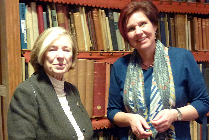 Eine ältere Dame links (Ursula Männle) mit einer elegant gekleideten Ruth Peetoom vor einem Bücherregal. 