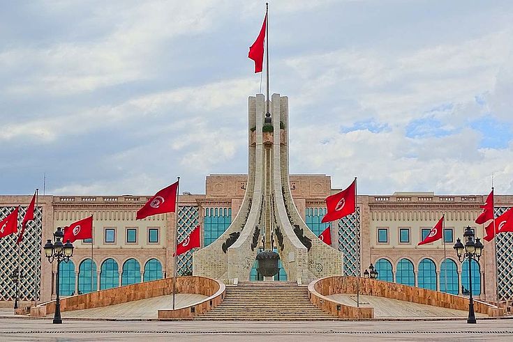 Ein hoher, nach unten martialisch breiter werdender Turm, den eine große tunesische Flagge ziert. Dahinter das repräsentative Parlamentsgebäude mit hohen Fensterfronten.