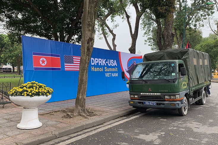 Es steht ein olivgrüner Lastwagen vor einem Gipfel-Plakat mit US- und Nordkorea-Flagge 