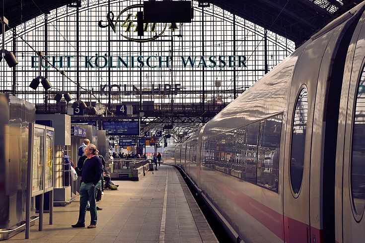 Der Bahnhof Köln. Am Gleis steht ein Mann und studiert den Fahrplan. Hinter ihm hält ein ICE, die Türen geöffnet.