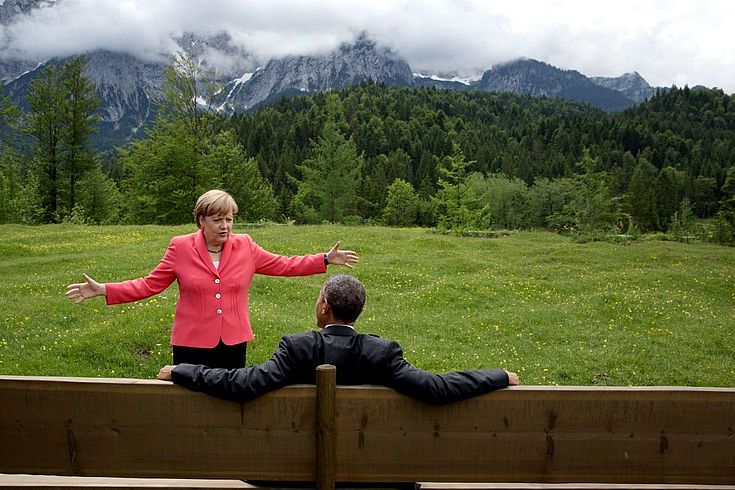 Obama sitzt raumfüllend in der Mitte einer Panoramabank vor Bergkulisse. Vor ihm steht mit ausgebreiteten Armen Angela Merkel und spricht intensiv auf den US-Präsidenten ein.