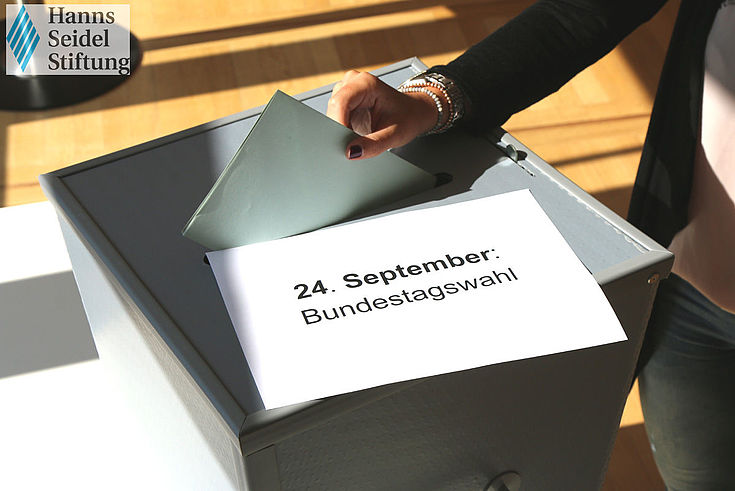 Wahlurne auf Schultisch. Hand wirft gefalteten Wahlzettel in Schlitz.