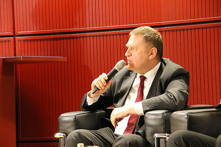 Huber sitzt im Anzug in einem Ledersessel auf dem Podium und spricht.