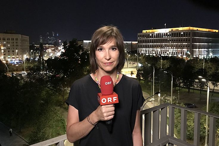 Carola Schneider steht mit einem Mikro in der Hand vor der nächtlichen Kulisse von Moskau.