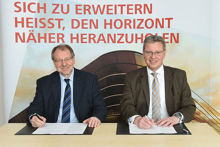 nwand, auf der stehDr. Peter Witterauf (HSS) und Dr. Roland Fleck sitzen lächelnd vor Unterschriftenmappen vor einer Leit: "Sich zu erweitern heißt, den Horizont heranzuholen"