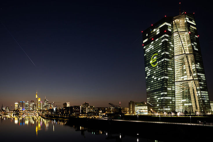 Das erleuchtete EZB-Gebäude am Fluss in Frankfurt. Ein großes Eurozeichen an der Glasfassade. 