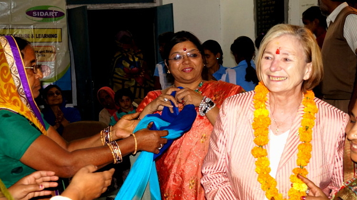 Ursula Männle (Mitte) beim Dorfparlament in Jaipur. Prof. Männle mit Blumenkette und indischer Kleidung wird von einer Gruppe indischer Frauen in Saris empfangen.
