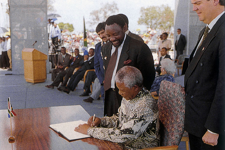 Nelson Mandela sitzt an einem Tisch und schreibt etwas auf ein Papier. Hinter ihm lächelnde Menschen.