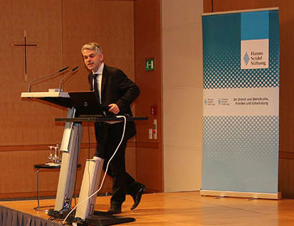 Masala am Rednerpult. Im Hintergrund HSS-Stellwand mit Logo.