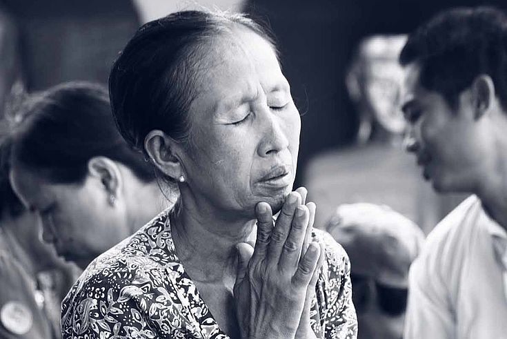 EIne ältere Frau sitz mit zum beten gefaltenen Händen auf dem Boden. Ihre Augen sind geschlossen. Ihr Ausdruck ist traurig bis hoffnungsvoll.