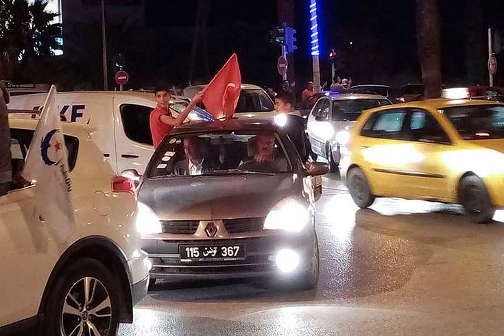 Es ist abends auf einer Straße in Tunis. Mehrere Autos, Fahrer schwenken die Flagge von Tunesien