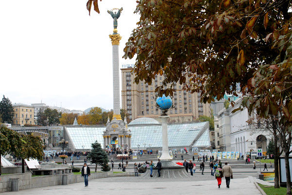 Teilansicht des Kiewer Maidan am Wahltag mit einem Baum im Vordergrund, einer Säule sowie Gebäuden im Hintergrund und einigen Menschen