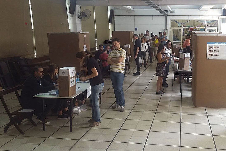Ein Wahllokal  mit einigen Personen in Buenos Aires. Zu sehen sind Wahlkabinen und ein Tisch zur Registrierung