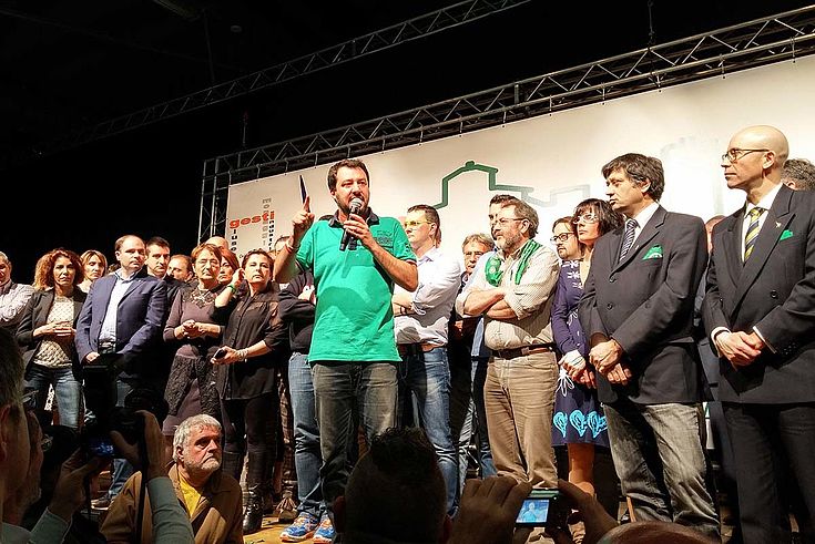 Salvini steht auf einer Bühne, umgeben von lauschenden Menschen und spricht offenbar von sich selbst überzeugt in ein Mikro.