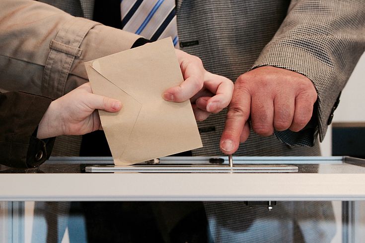 Eine Hand mit einem Stimmzettel über der Wahlurne, eine andere deutet auf den Schlitz.