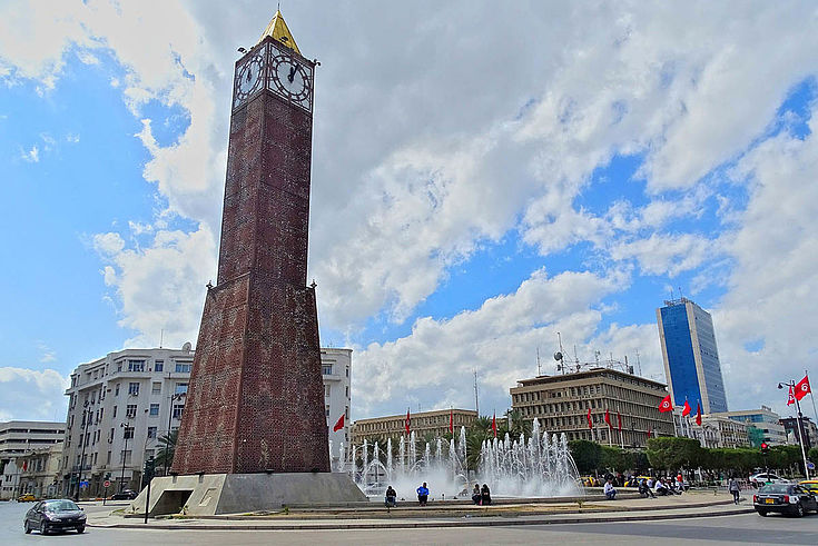 Ein großer Uhrenturm auf einem zentralen Platz in Tunis. Tunesische Flaggen im Halbrund im Hintergrund