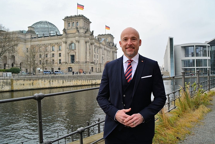 Vor einem Seitenarm der Spree steht Alexander Hoffmann, MdB, und lächelt in die Kamera. Im Hintergrund der Bundestag.