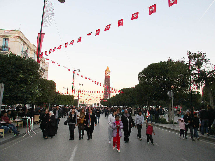 Eine mit Flaggen geschmückte Hauptstadtstraße, auf der Menschen in kleinen Grüppchen auf die Kamera zuschlendern. Im Hintergrund ein von der letzten Abendsonne beschienener Turm.