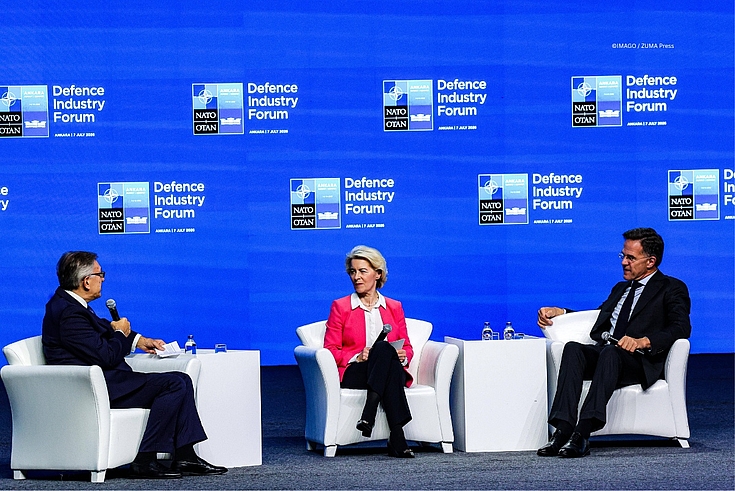 Ein Podium mit den Farben und dem Logo der Nato und des Verteidigungsindustrieforums. Drei Podiumsteilnehmer sitzten und sprechen miteinander. 