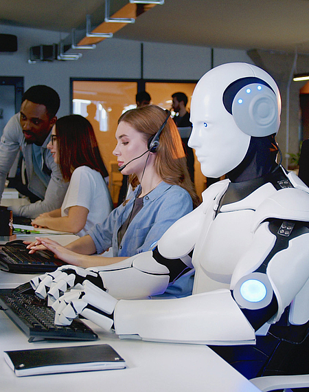 Ein Roboter arbeitet in einem Büro am Computer, daneben Personen am Computer