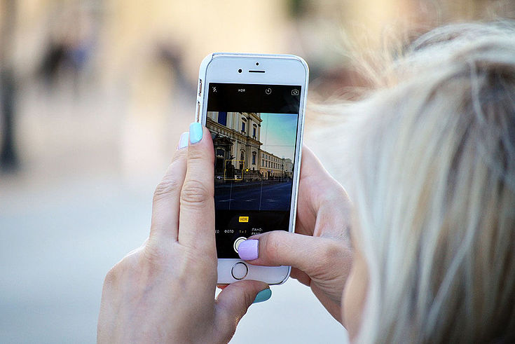 Frau hält Handy vor ein Gebäude und schießt ein Foto. Es ist auf dem Bildschirm abgebildet.