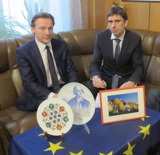 Die beiden Herren blicken ernst in die Kamera, an einem kleinen runden Tisch sitzend, über den eine Europaflagge gebreitet worden ist.