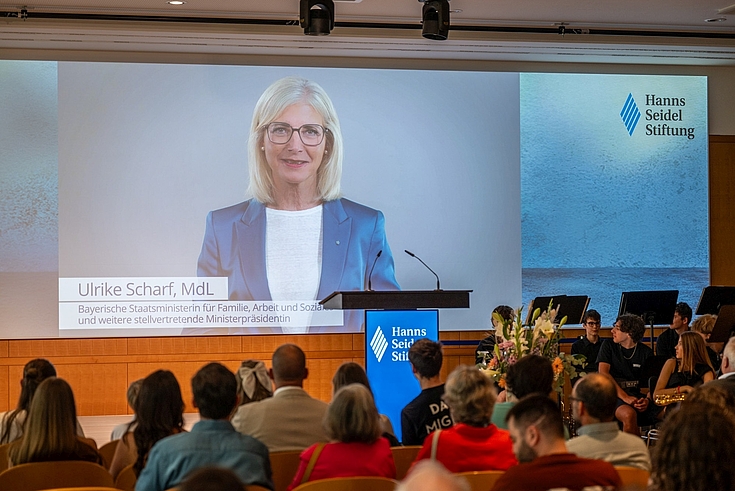 Staatsministerin Ulrike Scharf, MdL, wird auf dem Screen eingeblendet, davor Publikum