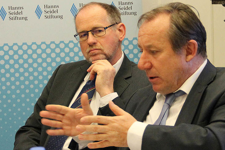 Am Verhandlungstisch: Vorderer Herr erklärt, hinterer Herr hört zu, die Hand am Kinn.