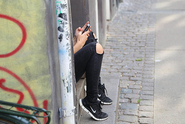 Die Beine eines jungen Menschens mit zerrissener Hose ragen aus einer Einbuchtung in einer Betonstruktur. Das Handy in der Hand ist auch zu sehen. Graffiti, Schmutz, Verwarlosung.