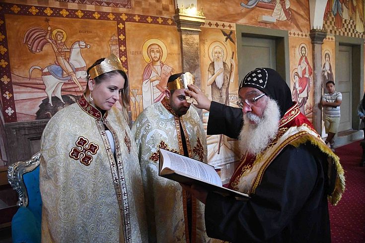 Der Bischof segnet eine junge Frau in prächtigem Ornat offenbar in einer orthodoxen Kirche mit alten Mosaiken an der Wand