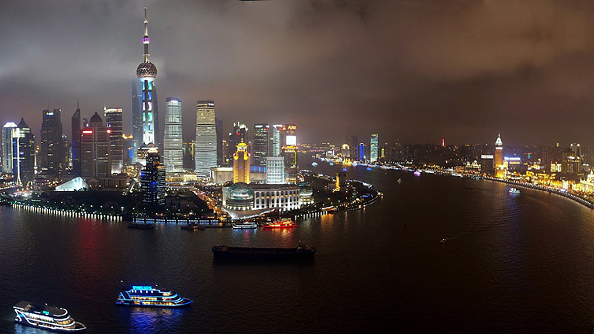 Hohe Wolkenkratzer, viel Glas, bei Nacht aus der Ferne aufgenommen. In einem weiten Halbkreis von tiefem Wasser umgeben.