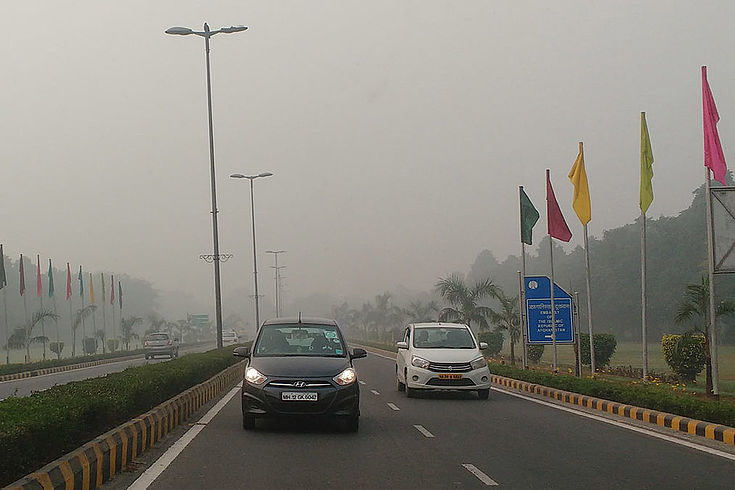 Breite Stadtstraße in Delhi mit Flaggen am rechten Rand, wo der Präsidentenpalast im dichten Smog nicht zu erkennen ist. Autos verschwinden im Dunst.