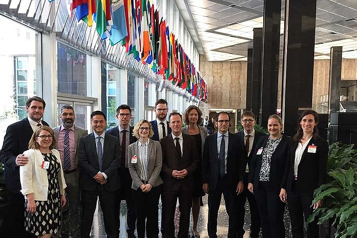 Gruppenbild in der Lobby eines größeren Gebäudes. Internationale Flaggen an der Decke, Pflanzen in großen Kübeln auf dem Marmorboden. Schick.