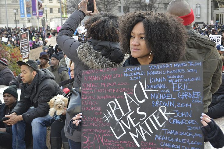 Eine junge schwarze Frau hält ein Plakat auf dem "Black Lives Matter" steht. Ruhiger, nachdenklicher Blick.