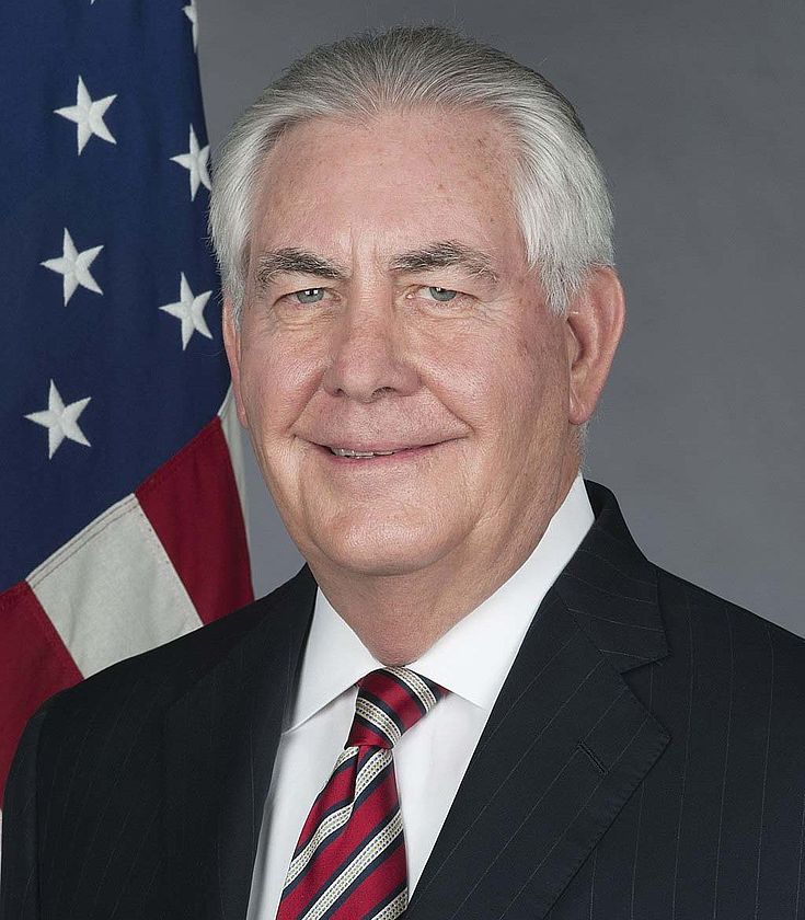 Ergrauter Mann in Anzug und Kravatte lächelt vornehm vor einer US-Flagge.